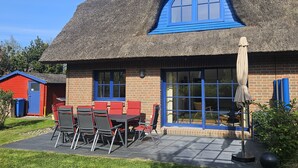 Een gebouw met rieten dak, een blauw omlijst raam, een terras met tafel en stoelen, en een kleine tuin.