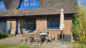 A thatched-roof building with blue-framed windows, a brick facade, and a patio with outdoor furniture. - Modern and comfortable vacation home - Reethaus Wiek left half of the house (Wiek)