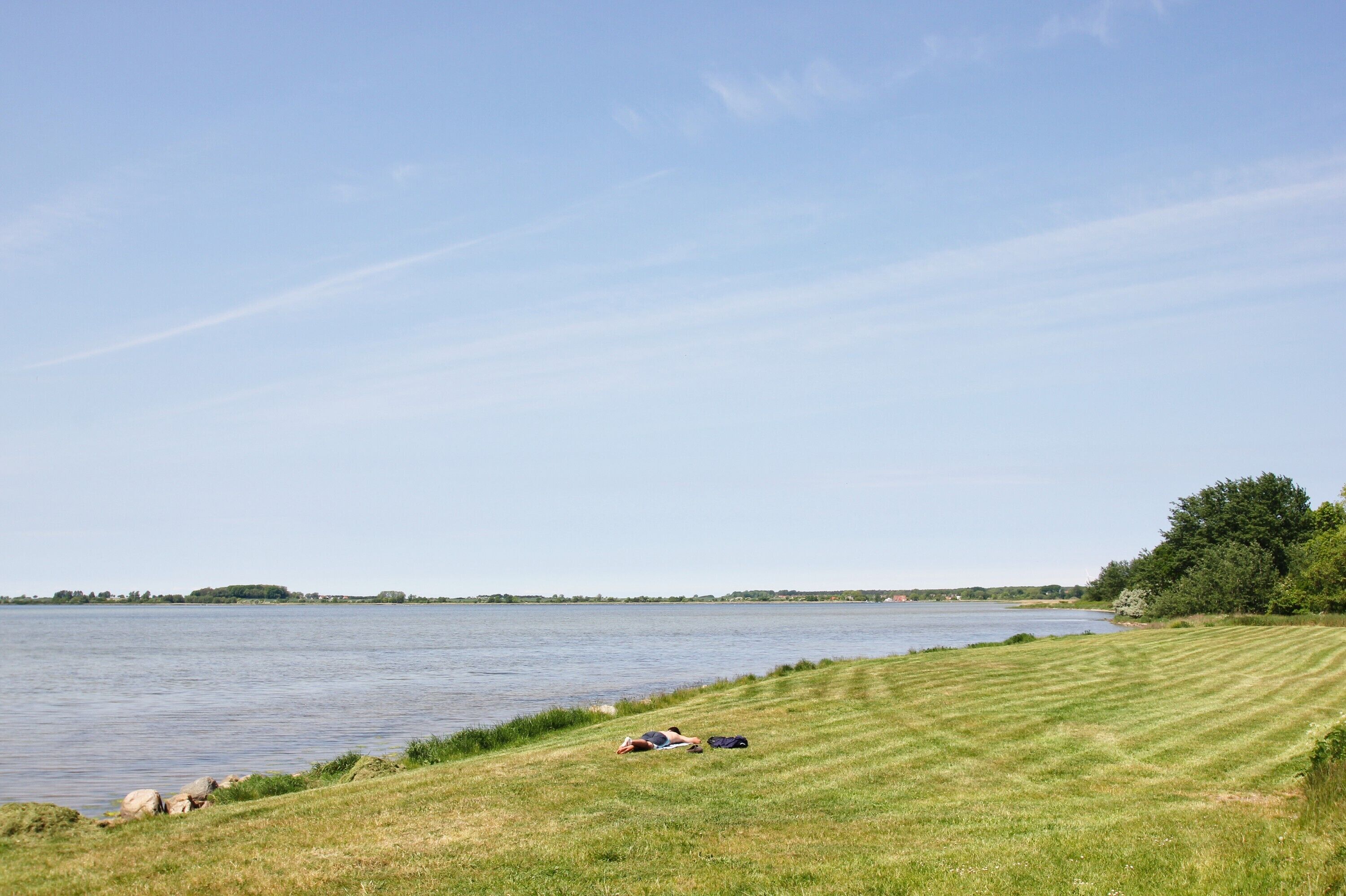 A grassy lakeside area with a person lying on the grass, a bag, and a small boat.