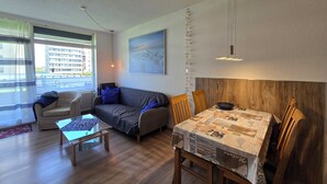 Ett modernt vardagsrum med en soffa, ett soffbord i glas, ett matbord med stolar och ett stort fönster med utsikt över byggnader.