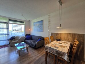 Ein modernes Wohnzimmer mit einer Couch, einem Glastisch, einem Esstisch mit Stühlen und einem großen Fenster mit Blick auf Gebäude.