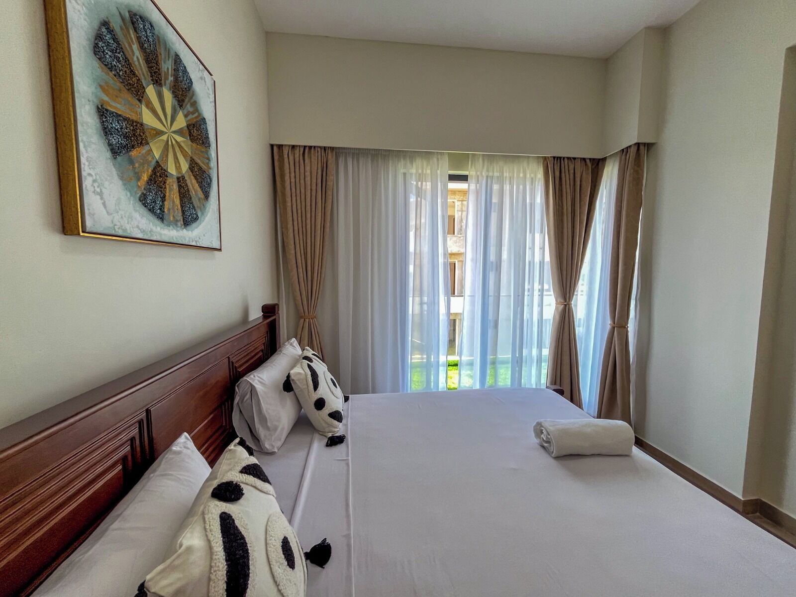 Una habitación de hotel con una cama grande, cabecera, una obra de arte colgada en la pared y vistas al exterior a través de cortinas traslúcidas.