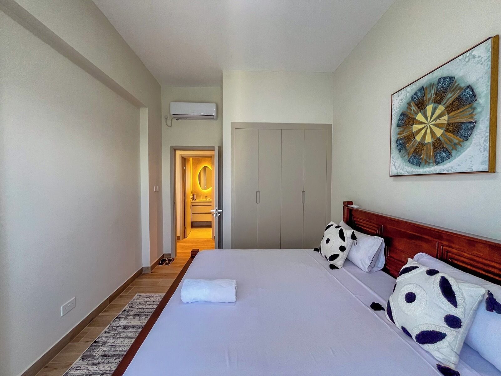 Una habitación de hotel con cama, cabecera de madera, una obra de arte en la pared, armario y aire acondicionado.