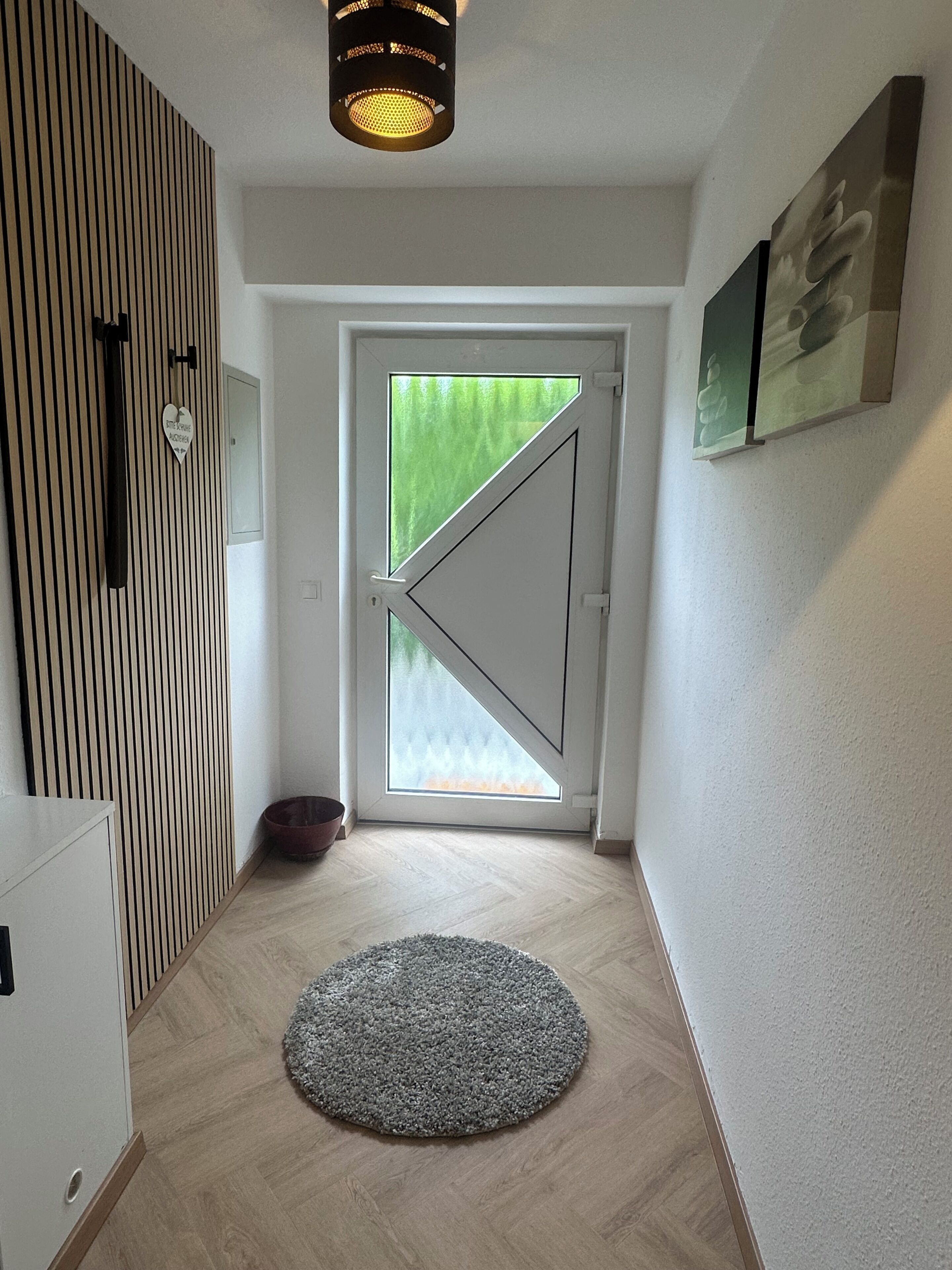 A hallway with a wooden slat wall, a round rug, a door with a triangular glass panel, and a ceiling light fixture.