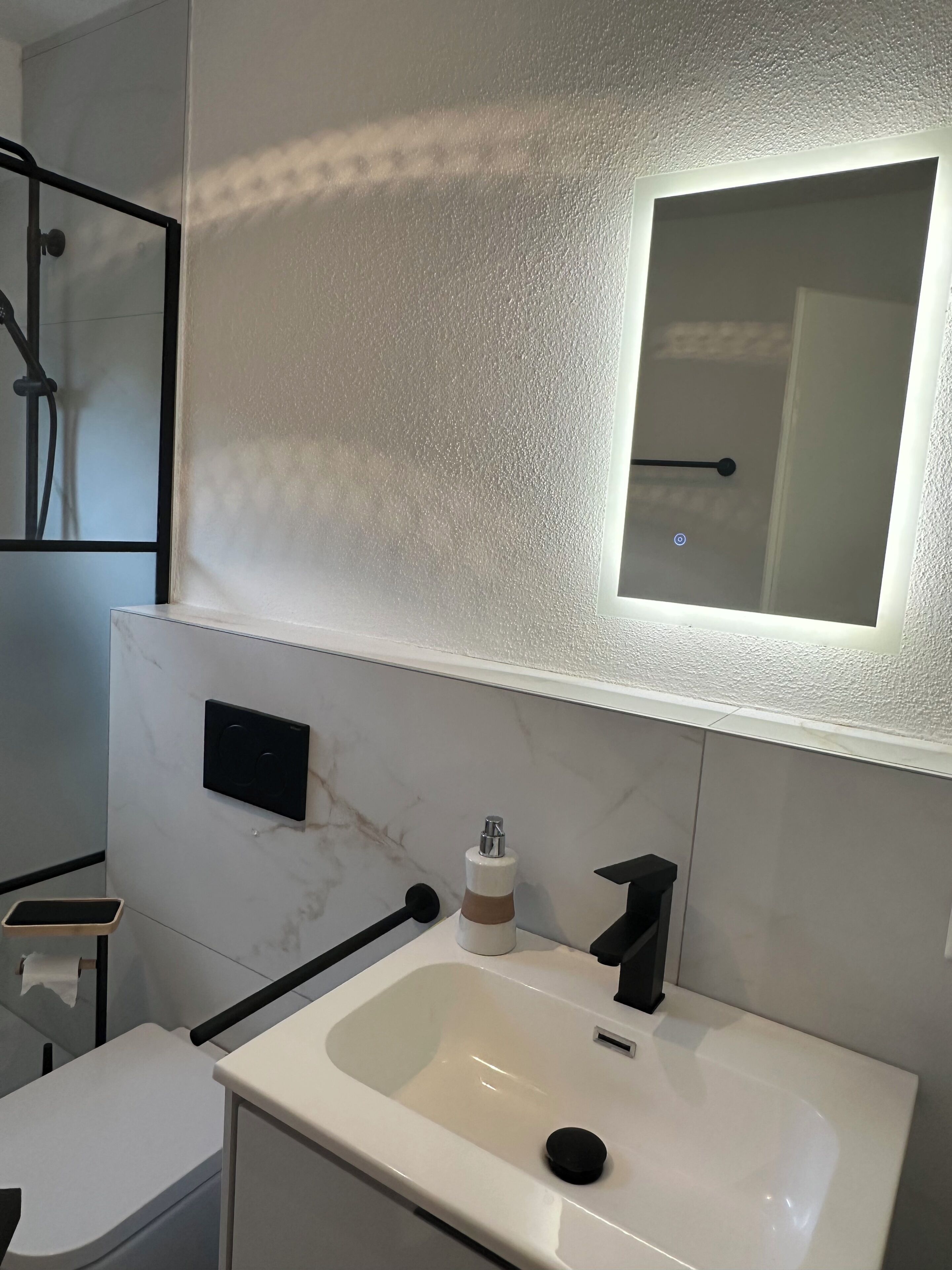 A modern bathroom with a white sink, a mirror, a towel dispenser, and a toilet.