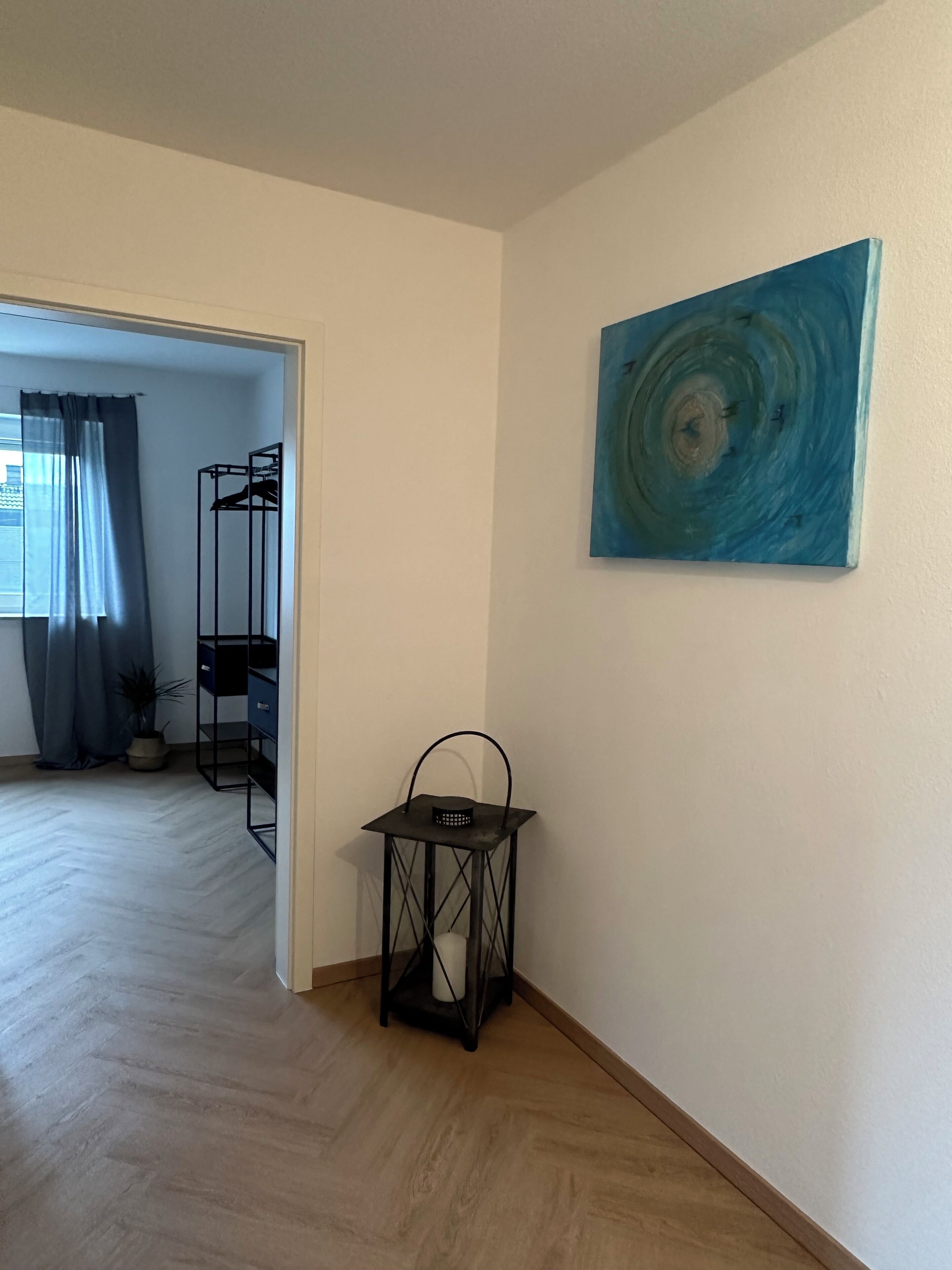 A room with a wooden floor, a black metal lantern, a blue abstract painting, a clothing rack, and a potted plant.