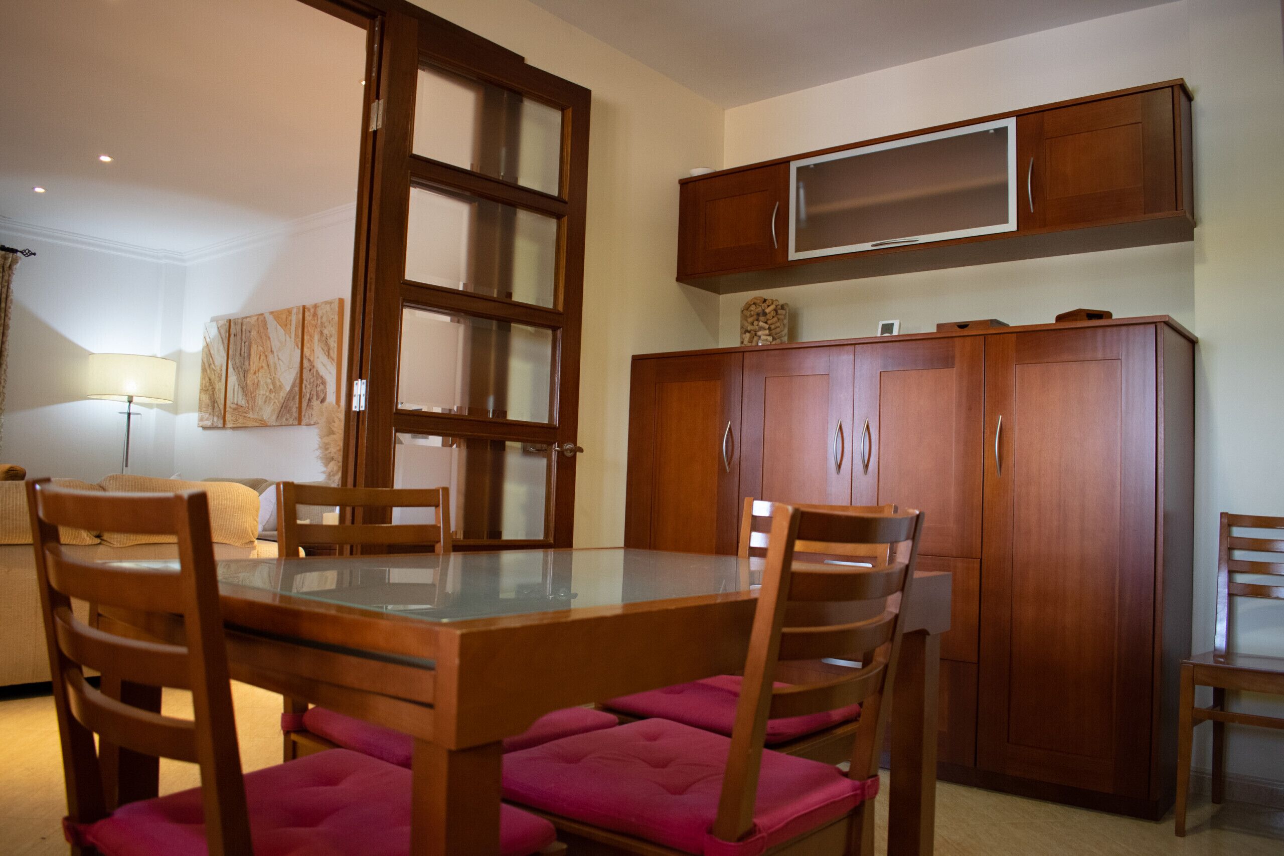 A dining area with a glass-top table, wooden chairs with cushioned seats, and a wooden cabinet with a television above.