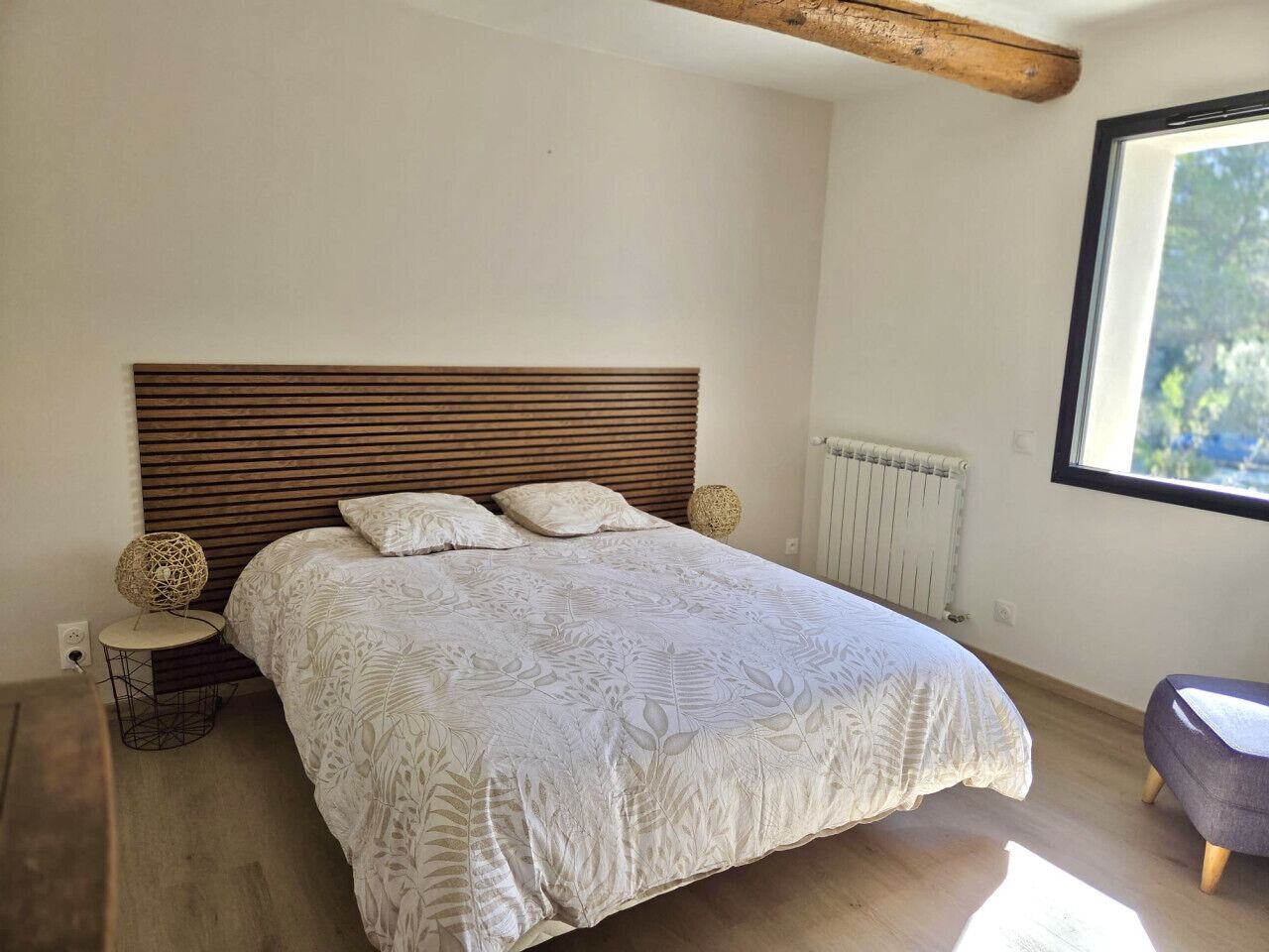 Un dormitorio con cabecera de madera, cama con colcha estampada, una mesita auxiliar redonda con un objeto decorativo y una ventana con vistas a follaje.