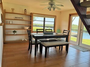Un espace repas comprenant une table, des chaises, un banc, un ventilateur de plafond, une fenêtre donnant sur l’extérieur et une étagère en bois agrémentée d’objets décoratifs.