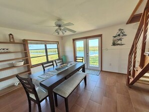 Zona pranzo con tavolo, sedie, ventilatore a soffitto e vista su un corpo d'acqua attraverso ampie finestre.