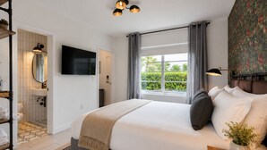 Un dormitorio con cama, televisión, ventana con cortinas, mesita de noche con planta y baño con espejo y lavamanos.