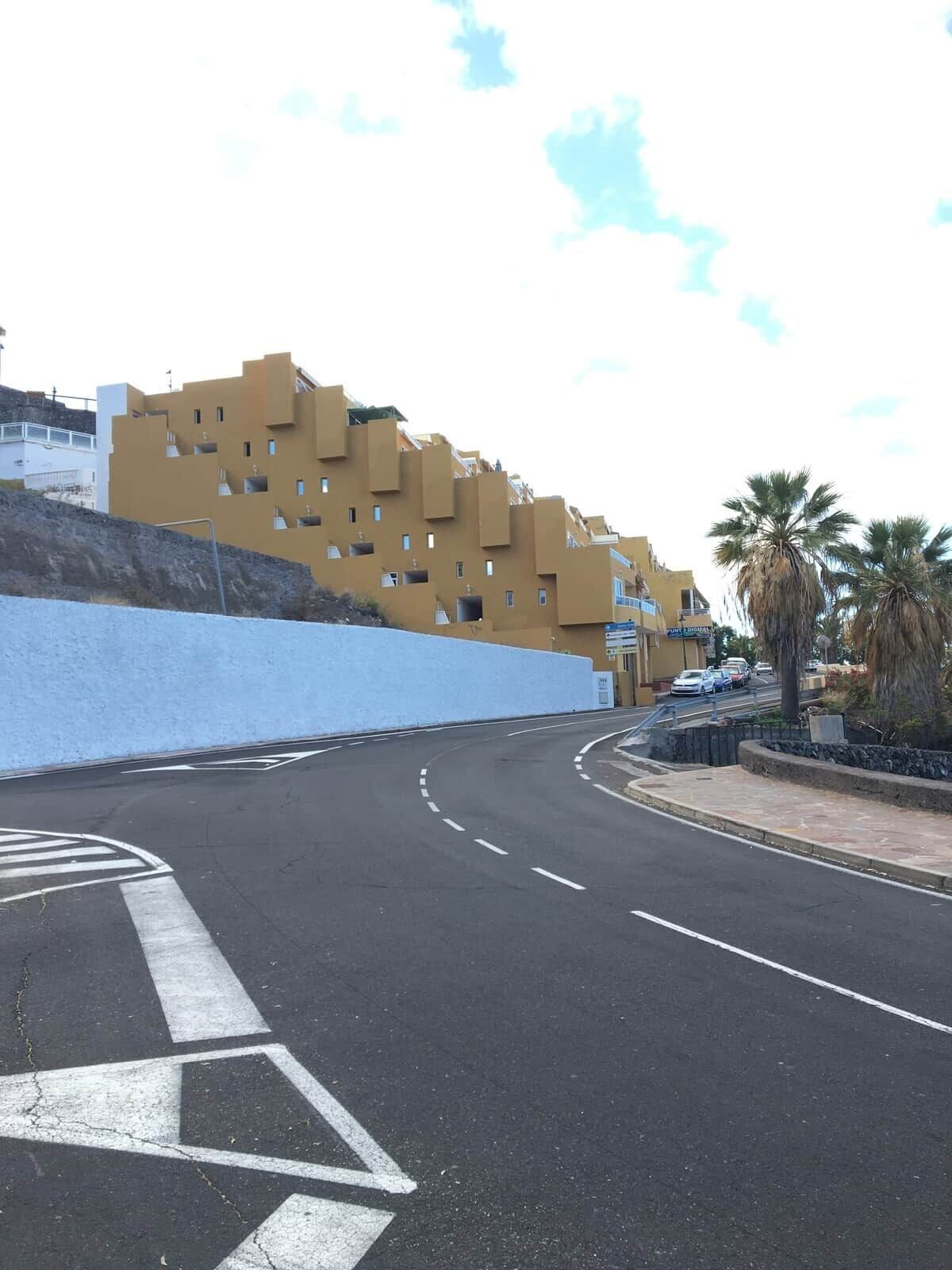 Une route sinueuse bordée d’une ligne blanche discontinue, un mur bleu et un immeuble jaune doté de nombreux balcons.