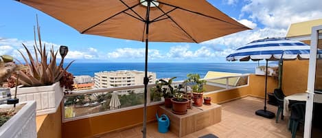 A balcony with a large umbrella, potted plants, a hanging chair, and a view of the sea and buildings.