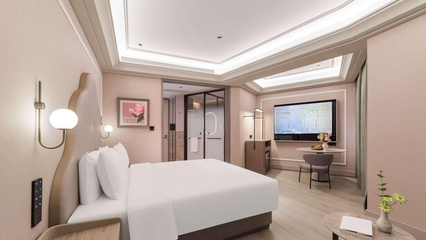 Una habitación de hotel moderna con una cama grande, mesita de noche, control remoto y un pequeño escritorio con una silla y un jarrón de flores.