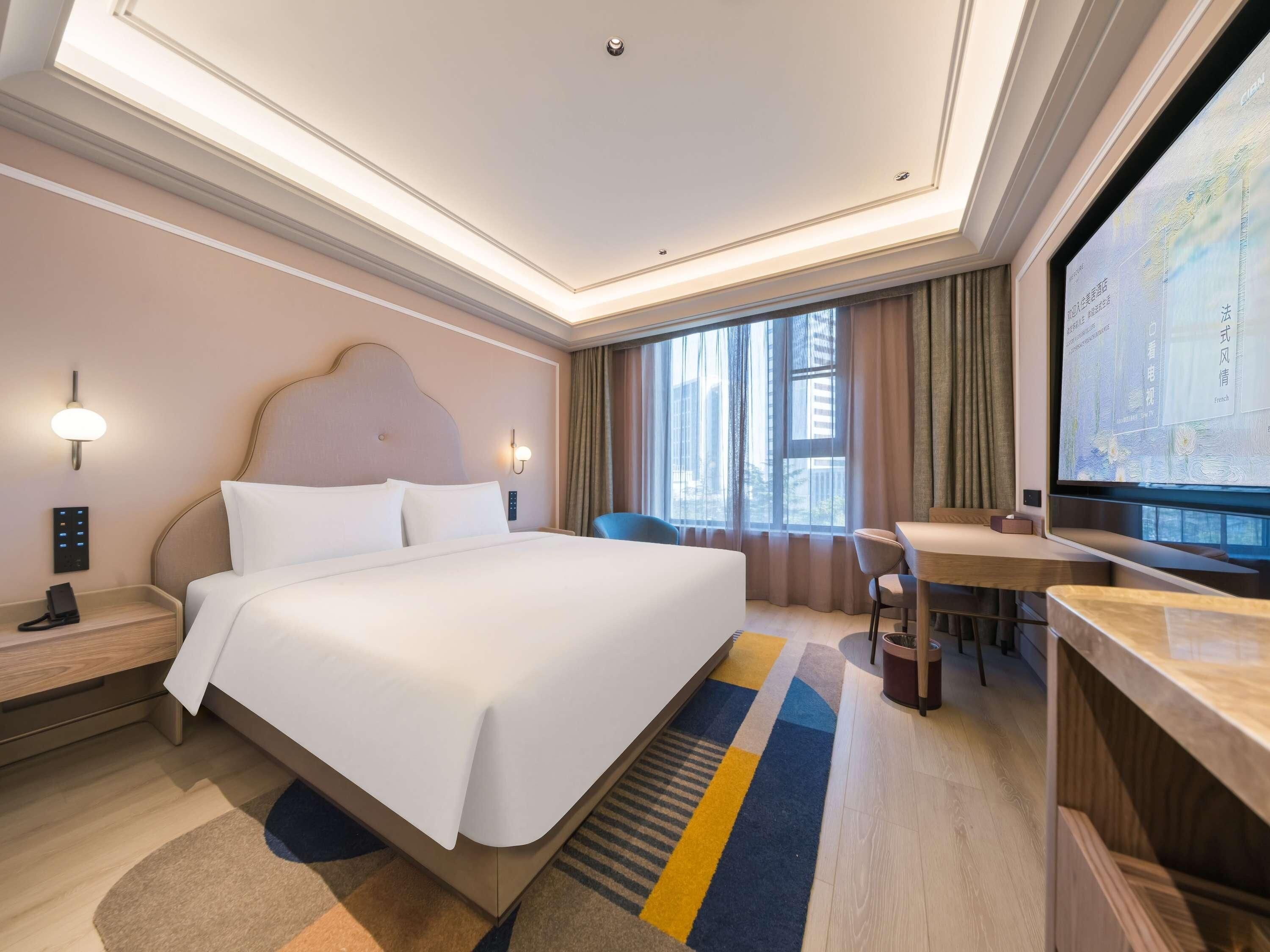 A hotel room with a large bed, bedside tables, a desk, a chair, a television, and a window with curtains.