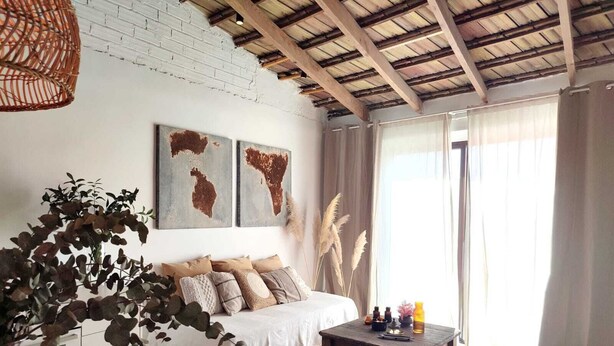 Ein Zimmer mit einem Bett, einem Holztisch, einer Pendelleuchte und einem großen Fenster mit Vorhängen.