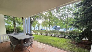 Un portico coperto con tavolo e sedie, un pavimento piastrellato e vista su una strada con auto parcheggiate e alberi.