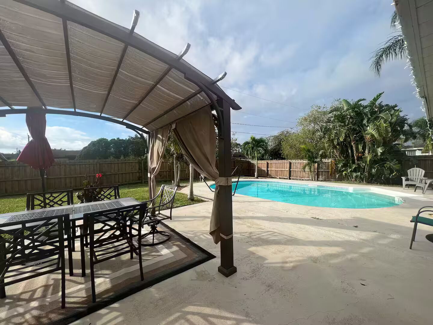 A covered patio area with a pool, outdoor furniture, and a wooden fence in the background.