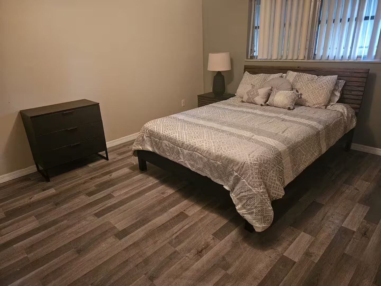 A bedroom with a bed, a dresser, a nightstand, a lamp, and a window with blinds.