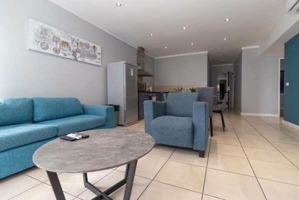Una sala de estar moderna con sofá, sillón y mesa de centro, una cocina al fondo y un pasillo que conduce a otra habitación.