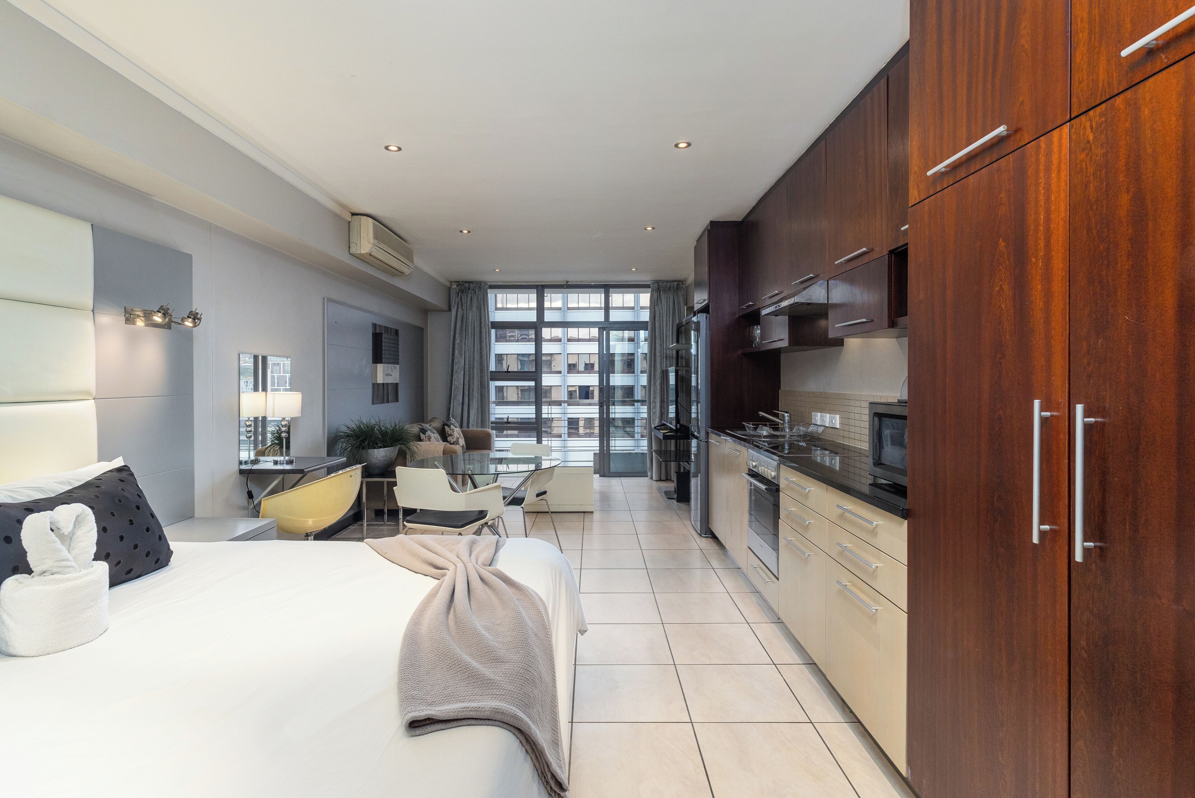 A modern hotel room with a bed, a desk, a chair, a kitchen area, and a view through large windows.