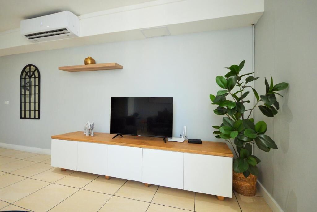 Een moderne woonkamer met een flatscreen televisie op een houten console, een wandplank, een plant in pot en een raam met decoratief roosterwerk.