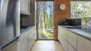 A modern kitchen with a stainless steel refrigerator, wooden cabinets, a built-in microwave, and a view of trees through a glass door.