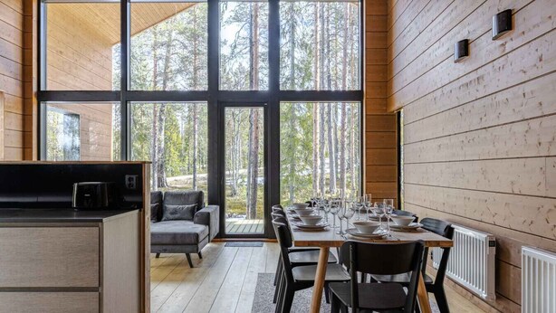 A modern kitchen with a dining area, large windows, and a view of the forest.