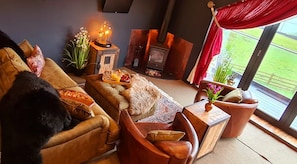 Ein gemütliches Wohnzimmer mit einer Couch, einem Beistelltisch mit einer Vase, einem Kamin und einem großen Fenster mit Blick auf Grünflächen.