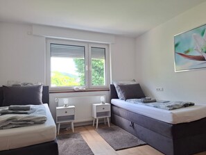 A bedroom with two beds, a window with blinds, a radiator, a small table, and a wall-mounted picture. - Apartment 'Melody' with Wi-Fi (Oberkochen)