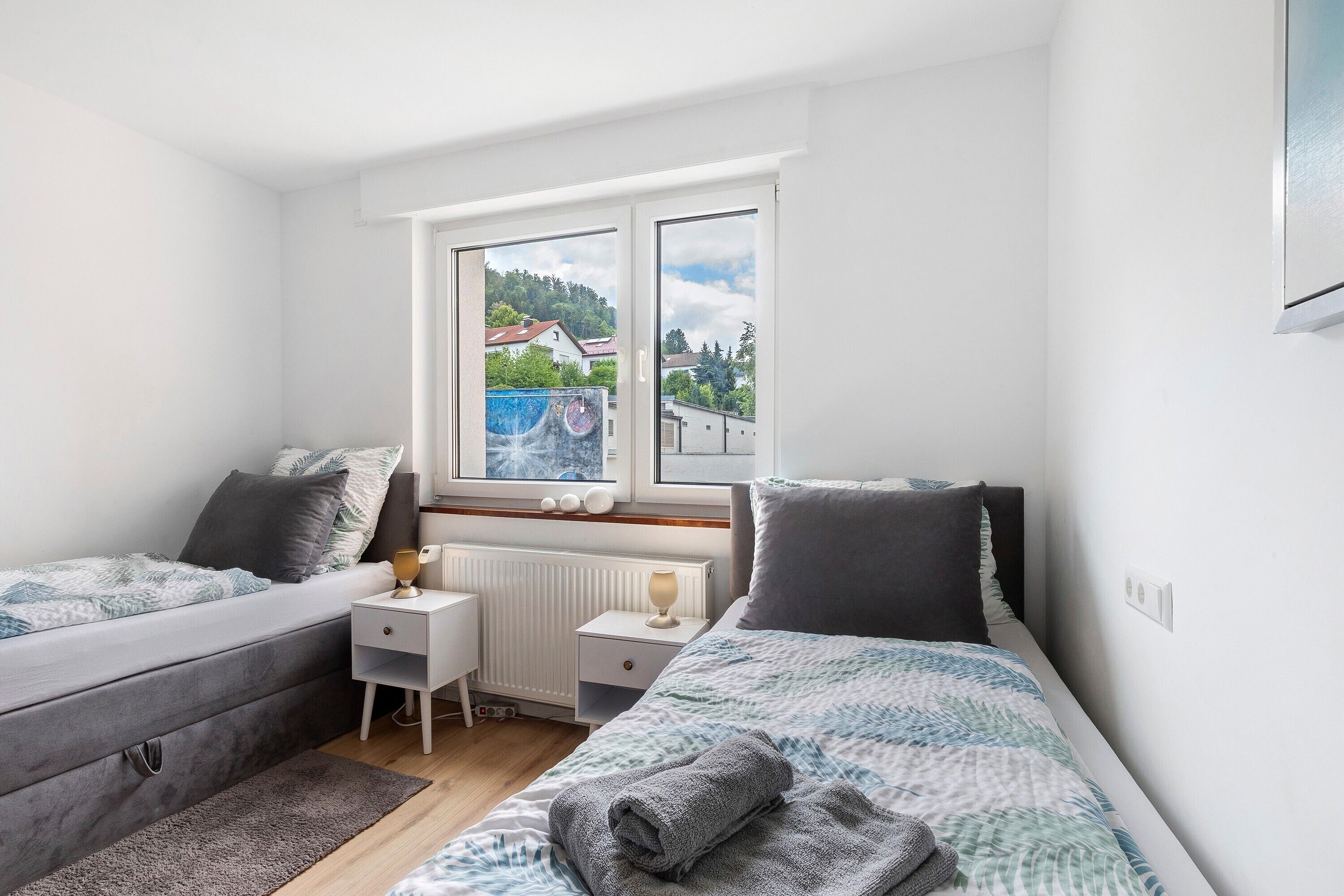 A bedroom with a bed, bedside tables, a radiator, and a window with a view of houses.