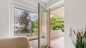 A modern living room with a sofa, a sliding glass door leading to an outdoor balcony, and a view of a residential area. - Apartment 'Melody' with Balcony and Wi-Fi (Oberkochen)