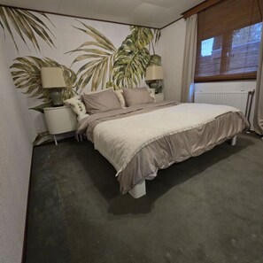 Une chambre à coucher comprenant un lit, deux lampes de chevet, une table de chevet, une grande fresque murale et une fenêtre avec des rideaux.