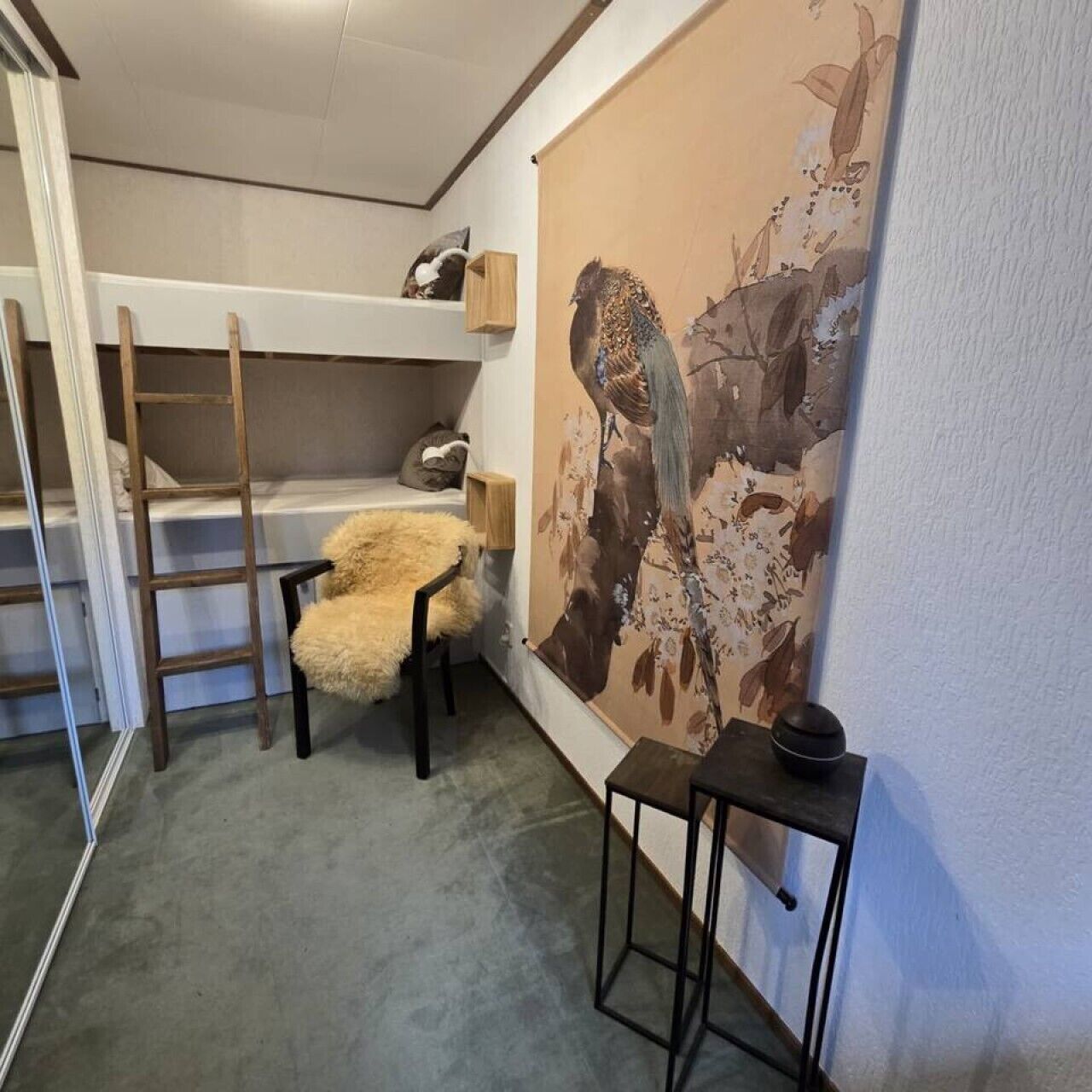 Huoneessa on kerrossänky, pehmeällä tekoturkiksella päällystetty tuoli, pieni pöytä ja suuri, kukkateemainen taulu seinällä.