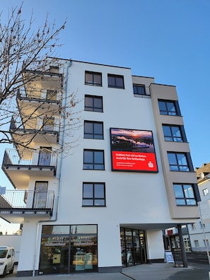 A multi-story building with a large digital billboard, a storefront with 'Amax' signage, and a tree in the foreground. - Modern city apartment with parking space on the doorstep (Koblenz)