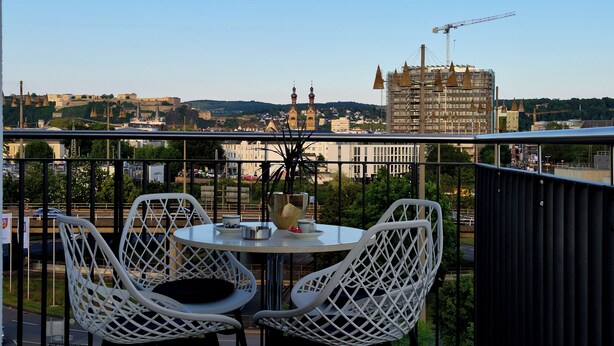 Un balcón con sillas blancas, una mesa y vistas de la ciudad, con edificios y una grúa de construcción.