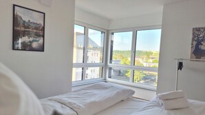 Un dormitorio con un ventanal grande, una cama con ropa de cama blanca y un cuadro en la pared.