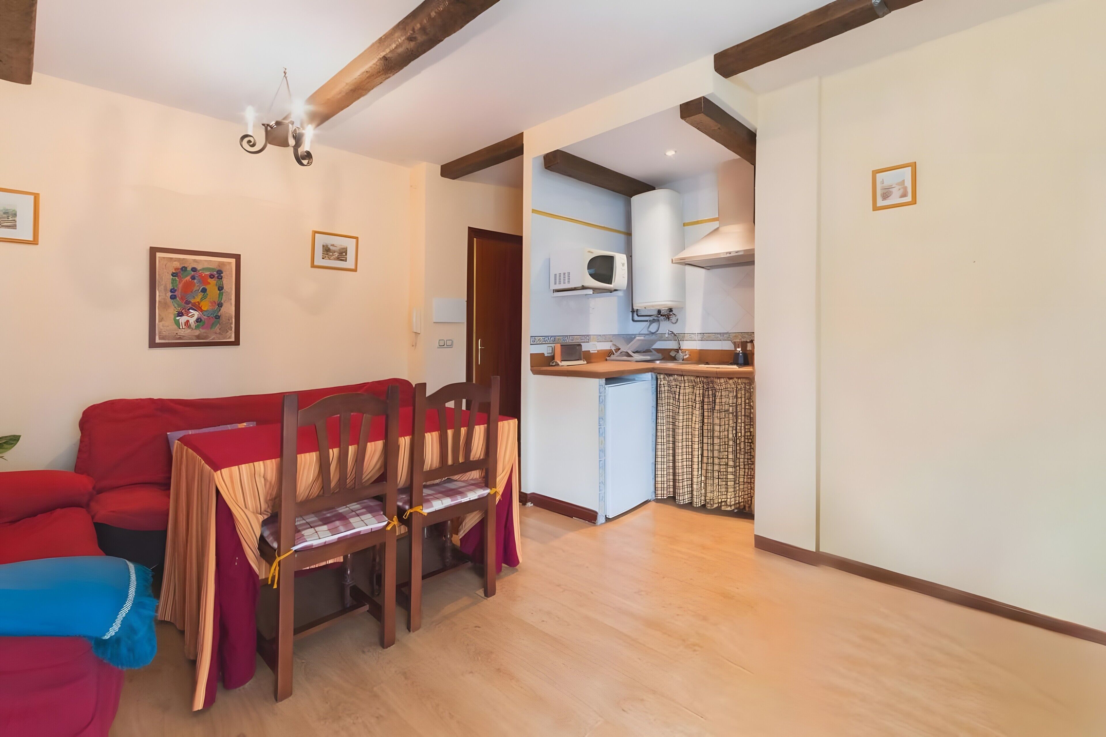 Una sala de estar con un sofá rojo, una mesa de comedor con sillas, una zona de cocina con microondas y un ventilador de techo.