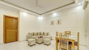 A modern living room with a sofa set, ottomans, a coffee table, and a wooden dining table with chairs. - Alaya Stays Zinnia 2BHK Apartment (Greater Noida)
