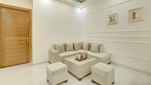 A modern living room with a sectional sofa, two ottomans, a coffee table, and a wooden door. - Alaya Stays Zinnia 2BHK Apartment (Greater Noida)