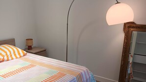 Een netjes opgemaakt bed met streepjesbeddengoed, een vloerlamp, een nachtkastje met een lamp en een spiegel aan de muur.