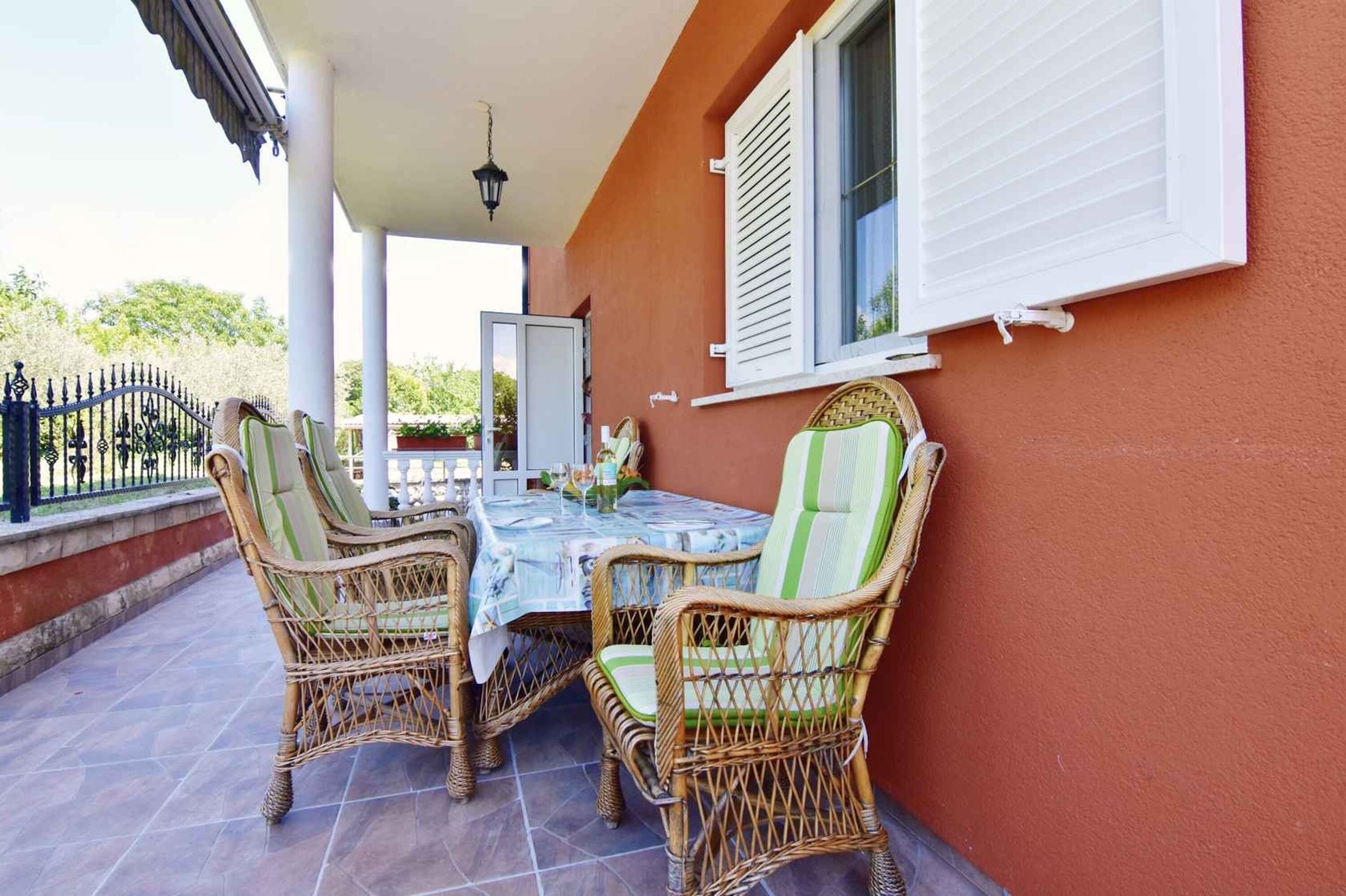 Une terrasse couverte, aménagée avec des meubles en osier, une table et une fenêtre blanche avec des volets.