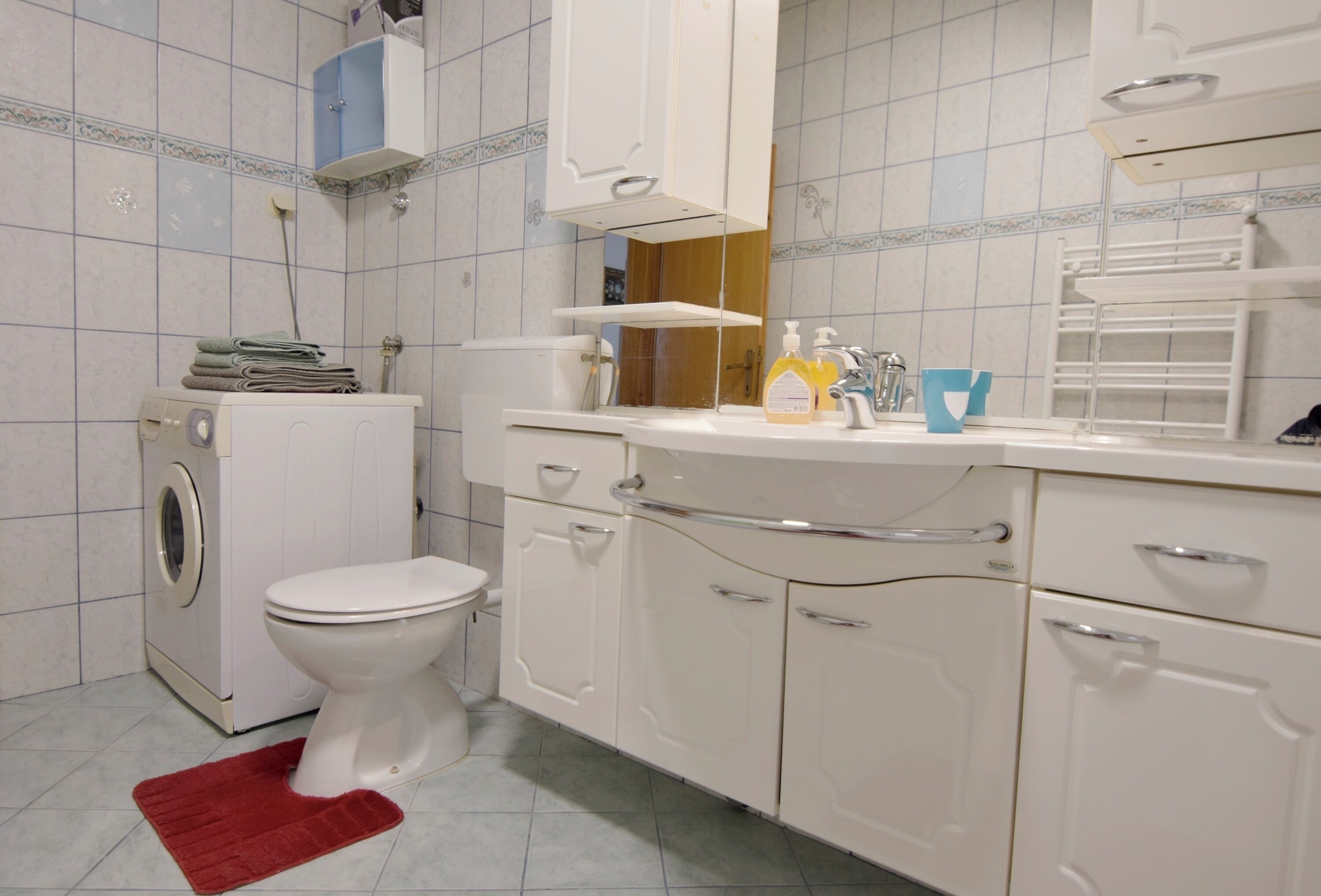 A bathroom with a toilet, washing machine, cabinets, and a mirror.