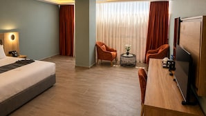 Une chambre d’hôtel comprenant un lit, deux fauteuils, un bureau avec un ordinateur, une petite table avec un vase et une grande fenêtre avec des rideaux.