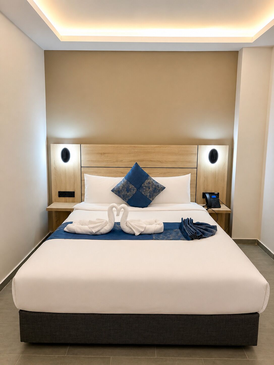 A hotel room with a large bed, two bedside tables with lamps, a wooden headboard, and a blue pillow.