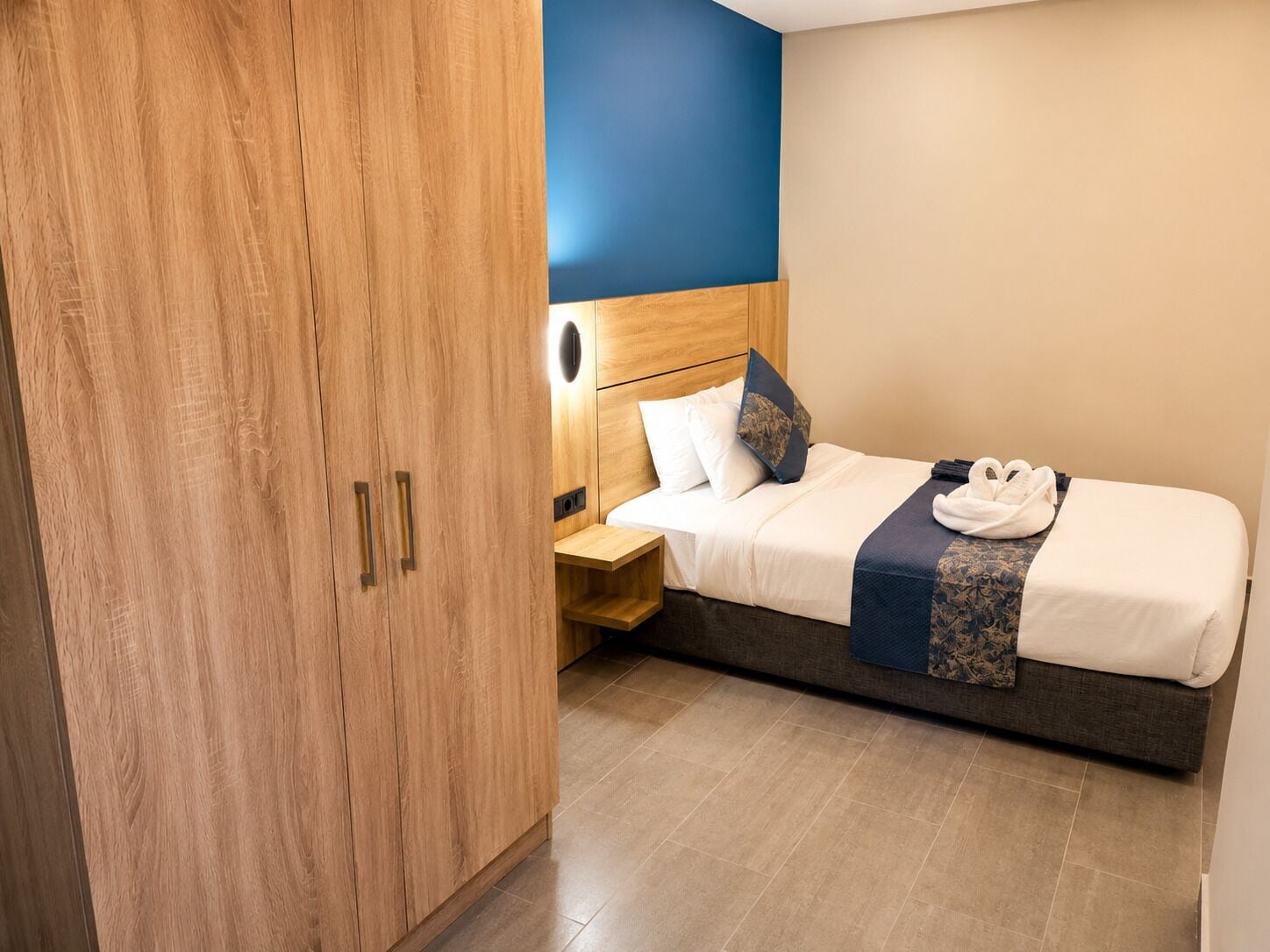 Habitación de hotel con un armario de madera, una cama con cabecero, una mesita de noche y una toalla sobre la cama.