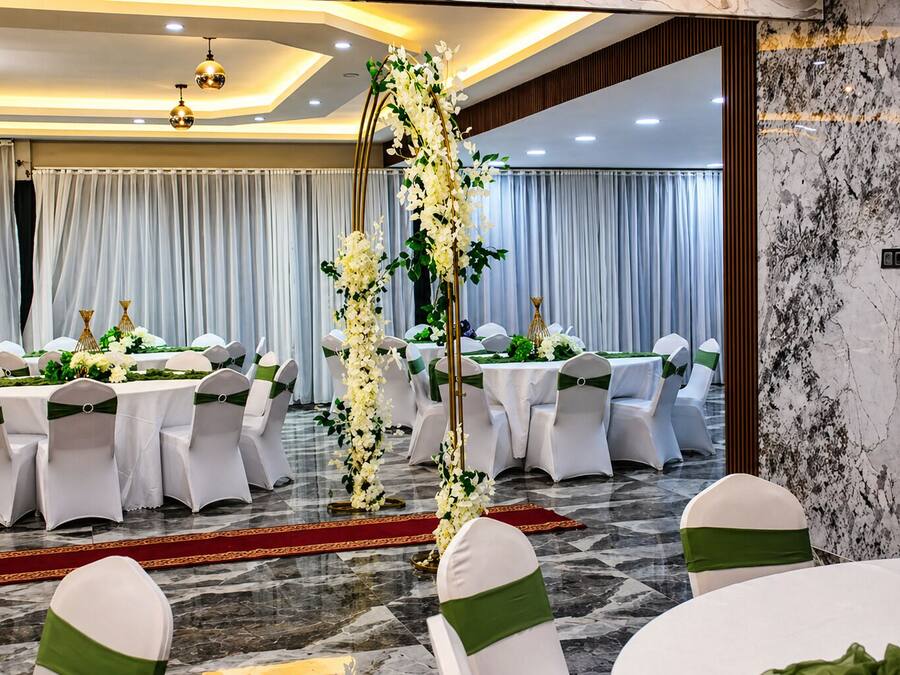 Un salón de banquetes con mesas redondas, sillas blancas con cintas verdes y un arco decorativo de flores.