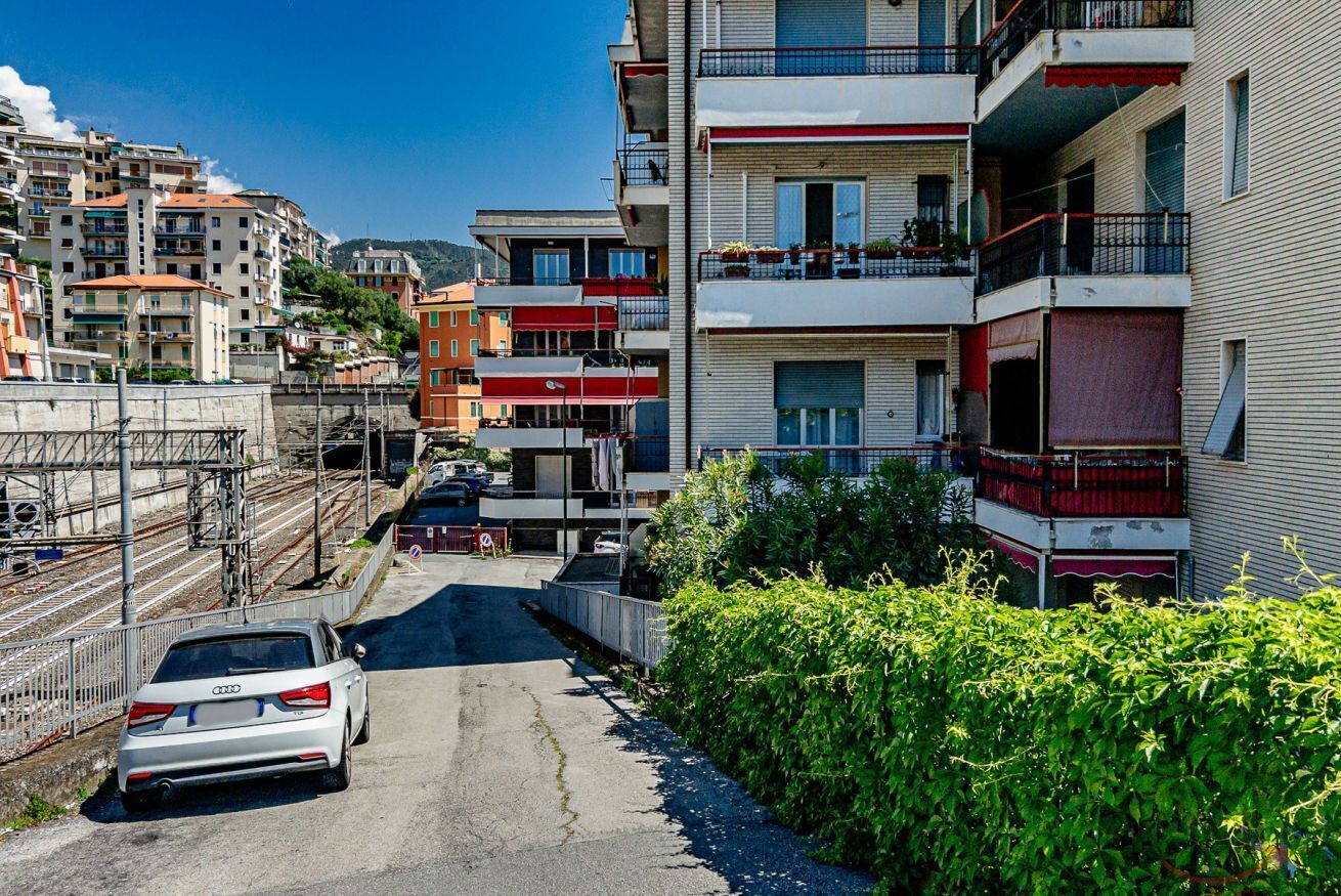 A narrow street with multi-story residential buildings, a parked car, and a hedge.