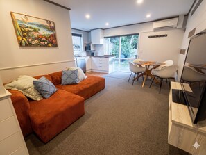 A modern living room with a sofa, a dining area with chairs, a kitchen in the background, and a large window.