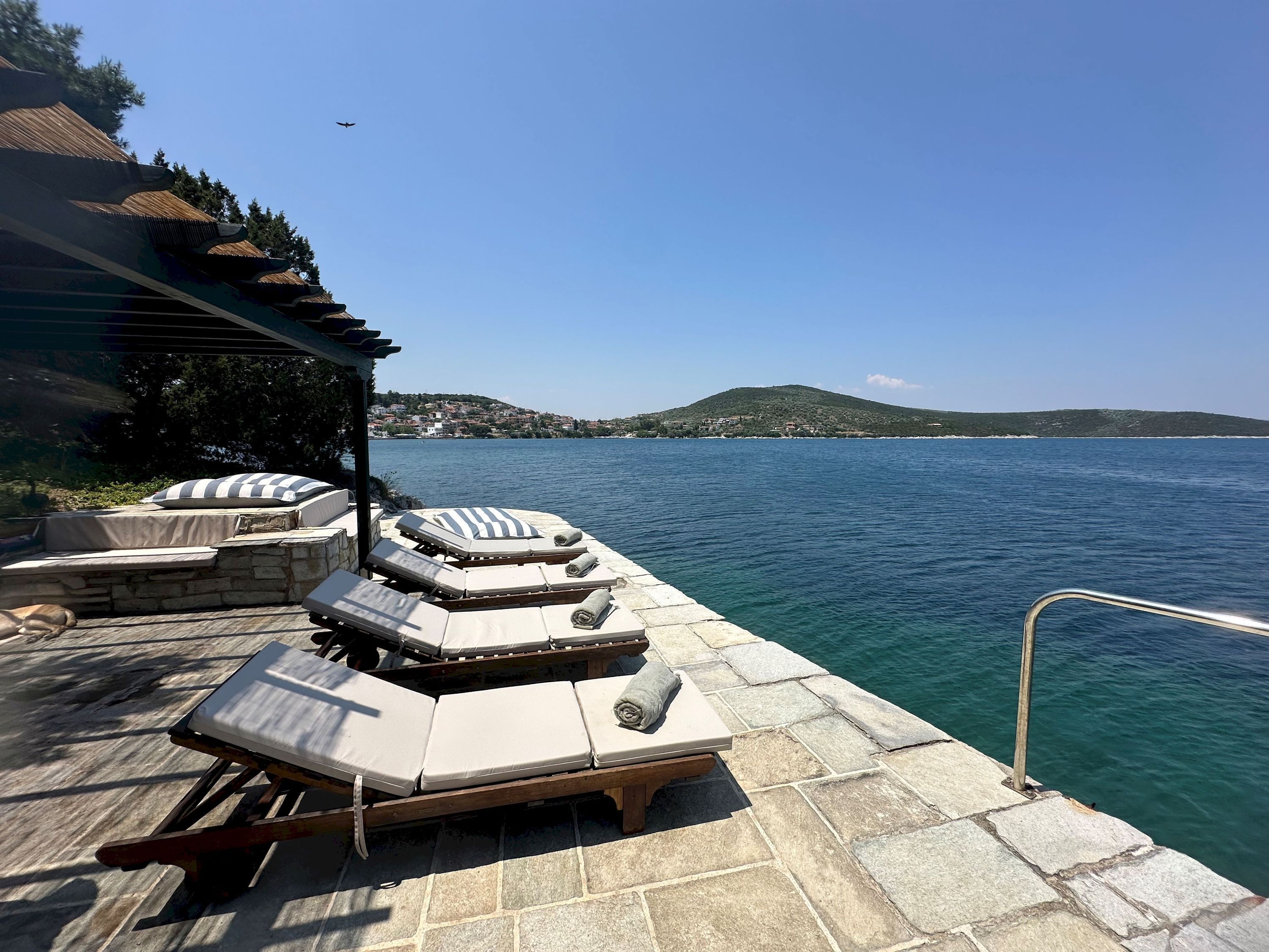 A stone deck with lounge chairs and a pergola overlooking a body of water.